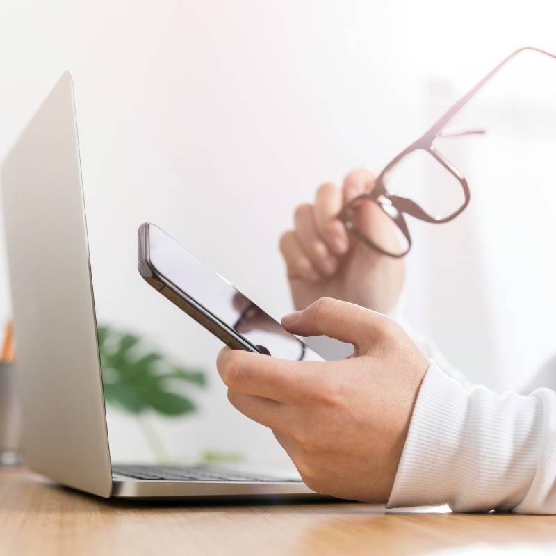 Hand holding eyeglasses beside a phone and computer